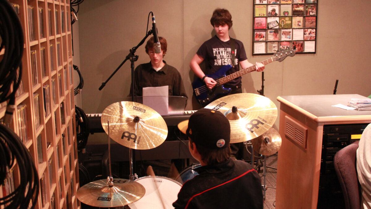 A youth playing a drum set, a youth playing a piano keyboard, and a youth playing an electric bass in a radio studio