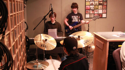 A youth playing a drum set, a youth playing a piano keyboard, and a youth playing an electric bass in a radio studio