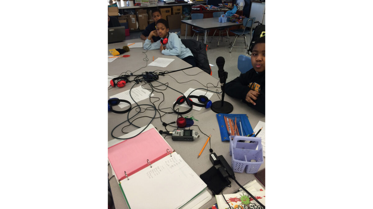 Three youth, one wearing headphones, sitting behind four sets of headphones, a portable recorder, and a microphone on classroom tables