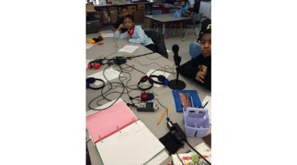 Three youth, one wearing headphones, sitting behind four sets of headphones, a portable recorder, and a microphone on classroom tables