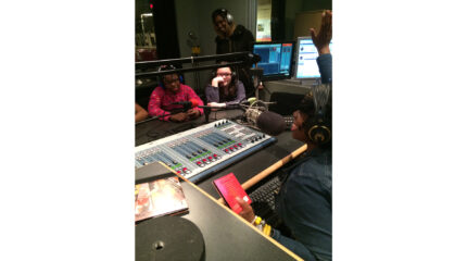 Two youth wearing headphones sitting behind microphones listening to another youth wearing headphones speaking into a microphone and sitting behind a mixer as another adult wearing headphones listens in the background in a radio studio