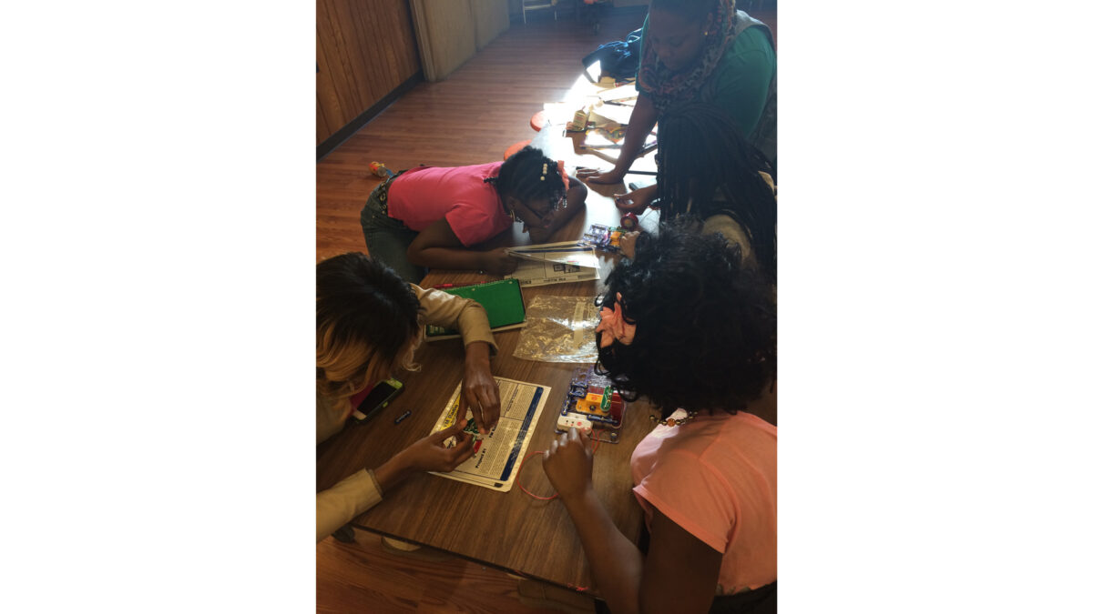 Four youth building electronic contraptions on a wooden classroom table