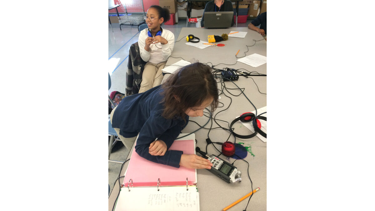 A youth wearing headphones looking off to the side and a youth interacting with a portable recorder on a classroom table