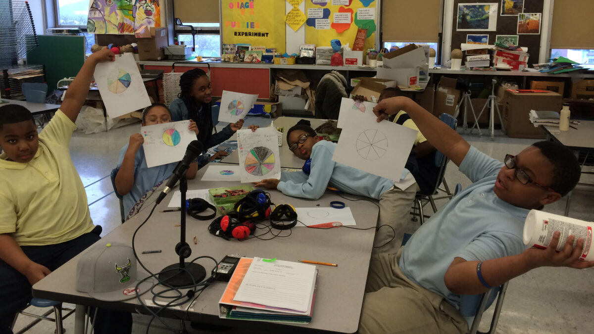 Five youth holding pie graphs on pieces of paper sitting around a microphone and six pairs of headphones on a classroom table