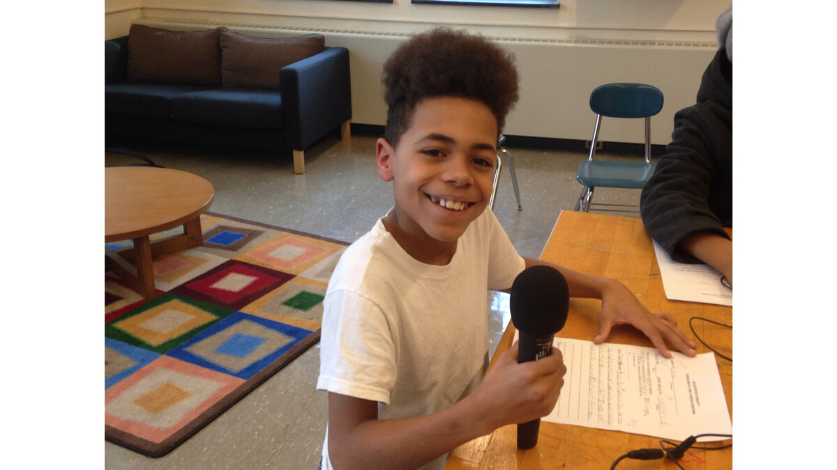 A youth smiling at the camera and holding a microphone while sitting behind a piece of paper on a wooden table