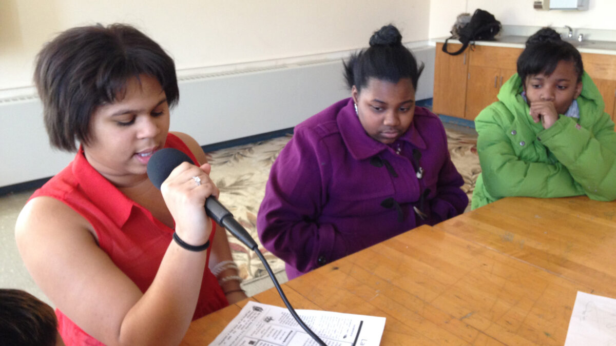 Two youth looking at another youth reading from a piece of paper into a microphone while sitting at a wooden table