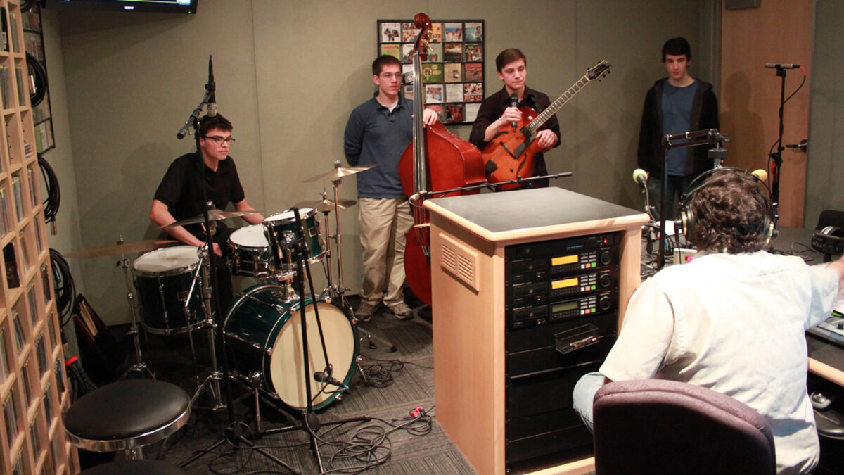 A teen sitting at a drumset, a teen holding an upright bass, a teen holding a guitar and speaking into a microphone, and a teen standing in the corner, all being interviewed by an adult wearing headphones in a radio studio