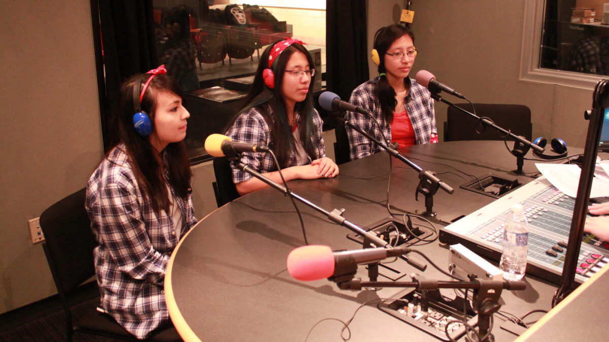 Three teens wearing headphones sitting behind microphones in a radio studio