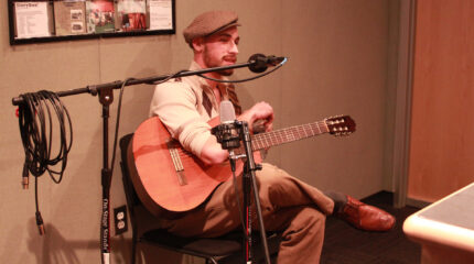 An adult sitting while holding an acoustic guitar and singing into a microphone in a radio studio