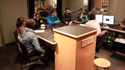 Six teens wearing headphones speaking into microphones being interviewed by an adult wearing headphones sitting behind a microphone in a radio studio