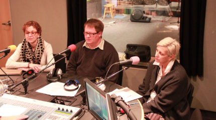 Three adults sitting behind microphones in a radio studio