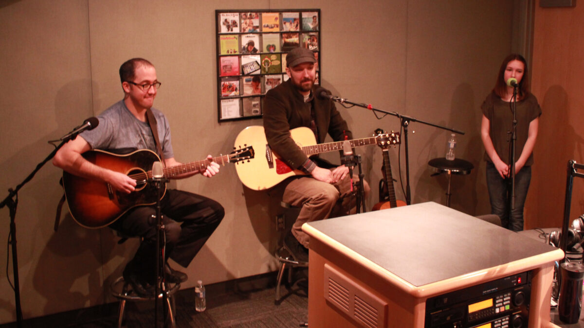 An adult playing acoustic guitar, an adult holding an acoustic guitar, and a teen singing into a microphone in a radio studio