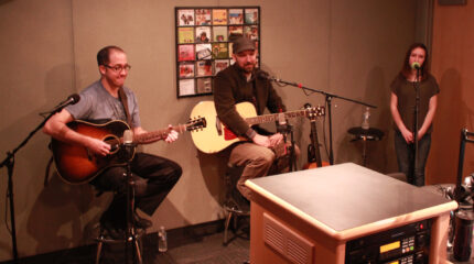 An adult playing acoustic guitar, an adult holding an acoustic guitar, and a teen singing into a microphone in a radio studio