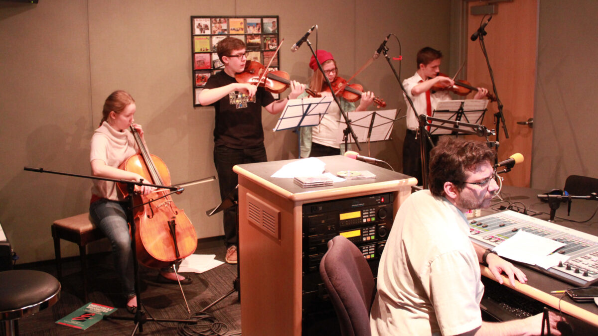 A teen playing cello, a teen playing viola, and two teens playing violin, all looking at sheet music on music stands in a radio studio