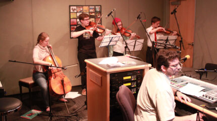 A teen playing cello, a teen playing viola, and two teens playing violin, all looking at sheet music on music stands in a radio studio