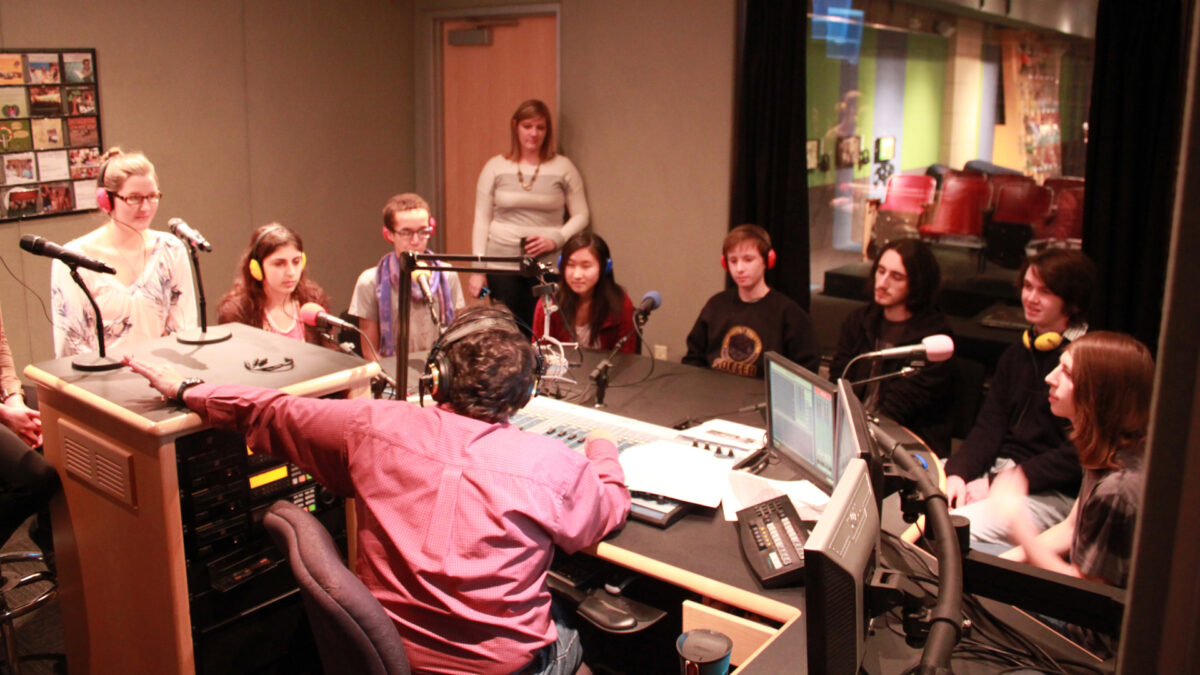 Eight teens wearing headphones speaking into microphones being interviewed by an adult wearing headphones sitting behind a microphone and a mixer as an adult listens in the background in a radio studio