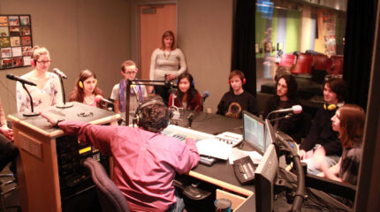 Eight teens wearing headphones speaking into microphones being interviewed by an adult wearing headphones sitting behind a microphone and a mixer as an adult listens in the background in a radio studio