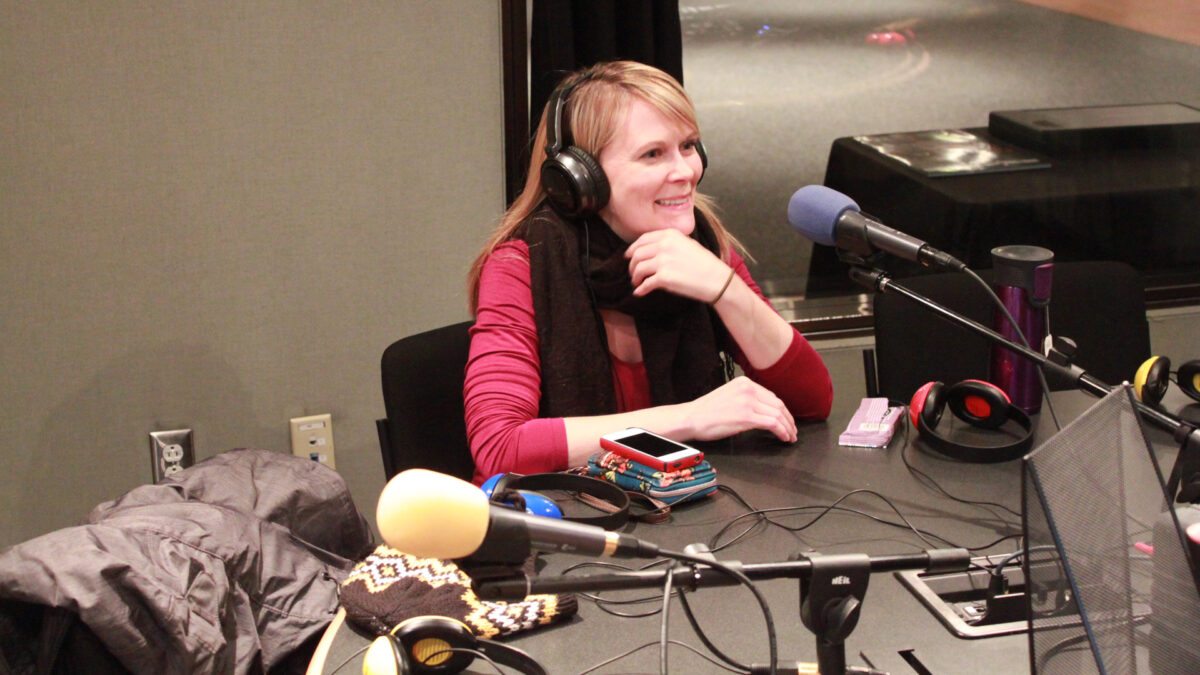 An adult wearing headphones sitting and smiling into a microphone in a radio studio