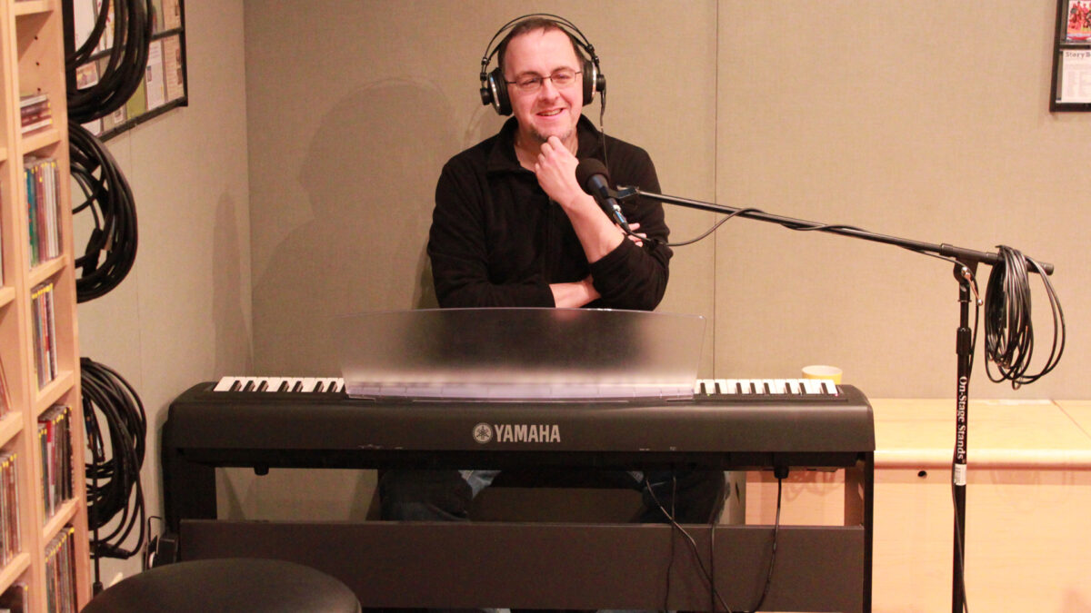 An adult wearing headphones smiling while sitting at a piano keyboard behind a microphone in a radio studio