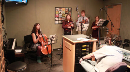A teen holding a cello and speaking into a held microphone, two teens holding a violin, and a teen holding a viola, all behind sheet music on music stands being interviewed by an adult wearing headphones and sitting behind a microphone and a mixing board in a radio studio