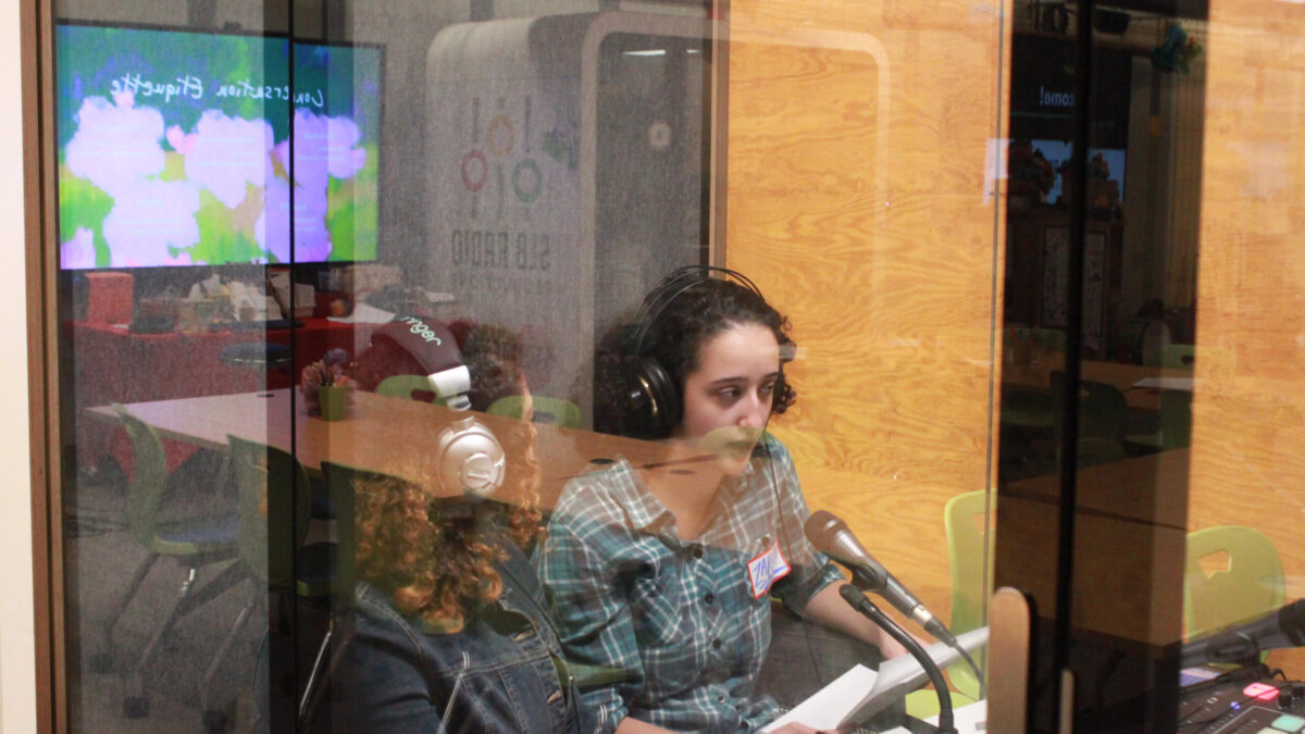 unnamed (1) Two teens wearing headphone speaking into a microphone in a recording pod