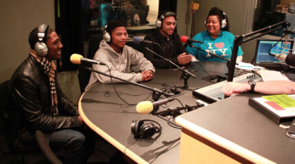 Four teens wearing headphones speaking into microphones in a radio studio