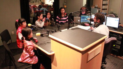 Five youth and an adult sitting and speaking into microphones being interviewed by an adult wearing headphones around their neck and sitting behind a microphone in a radio studio