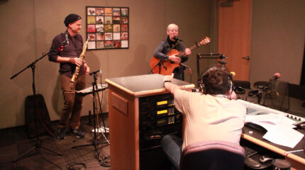 An adult holding a soprano sax and an adult holding an acoustic guitar speaking into a microphone being interviewed by an adult wearing headphones sitting behind a microphone and a mixer in a radio studio