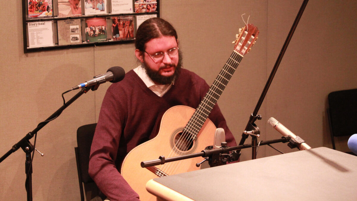An adult sitting while holding an acoustic guitar and speaking into a microphone in a radio studio