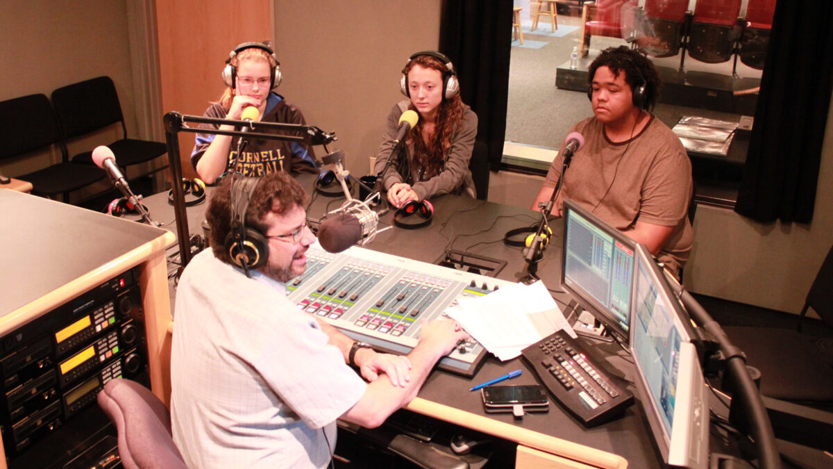 Three teens wearing headphones speaking into microphones being interviewed by an adult wearing headphones sitting behind a microphone and a mixing board in a radio studio