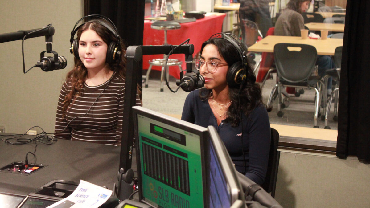 Two teens wearing headphone sitting behind microphones in a radio studio