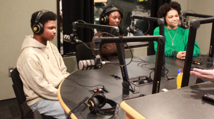Two teens and an adult wearing headphones sitting behind microphones in a radio studio