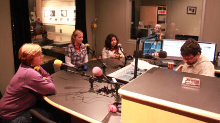 Two adults wearing headphones around their necks and a youth, all sitting behind microphones being interviewed by an adult sitting behind a microphone and a mixer in a radio studio