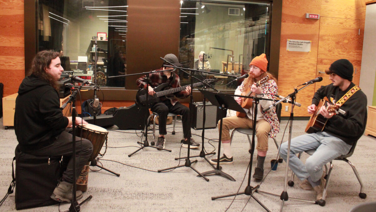 An adult playing a djembe, an adult playing an electric bass, and two adults playing acoustic guitars, all sitting behind microphones