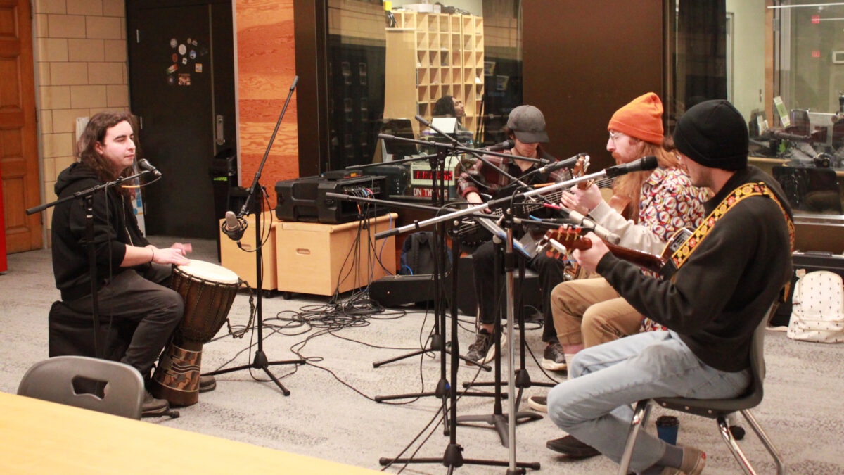 An adult playing a djembe, an adult playing an electric bass, and two adults playing acoustic guitars, all sitting behind microphones