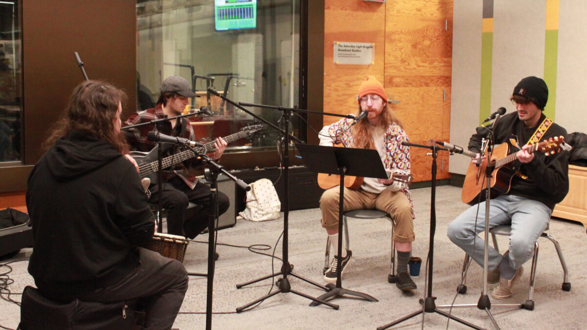 An adult playing a djembe, an adult playing an electric bass, and two adults playing acoustic guitars, all sitting behind microphones