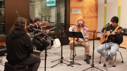 An adult playing a djembe, an adult playing an electric bass, and two adults playing acoustic guitars, all sitting behind microphones