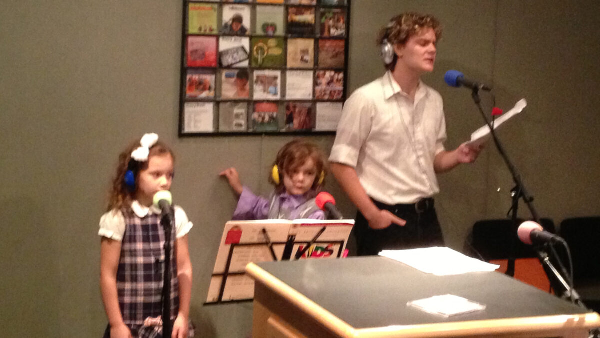 Two youth and a teen wearing headphones looking at sheet music and singing into microphones in a radio studio