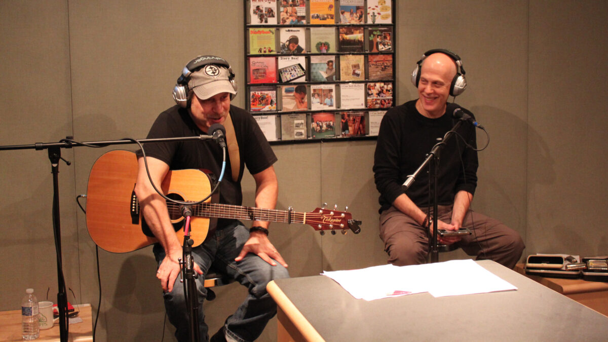 An adult wearing headphones sitting behind a microphone and holding a harmonica looking at an adult wearing headphones and holding an acoustic guitar while sitting and speaking into a microphone in a radio studio