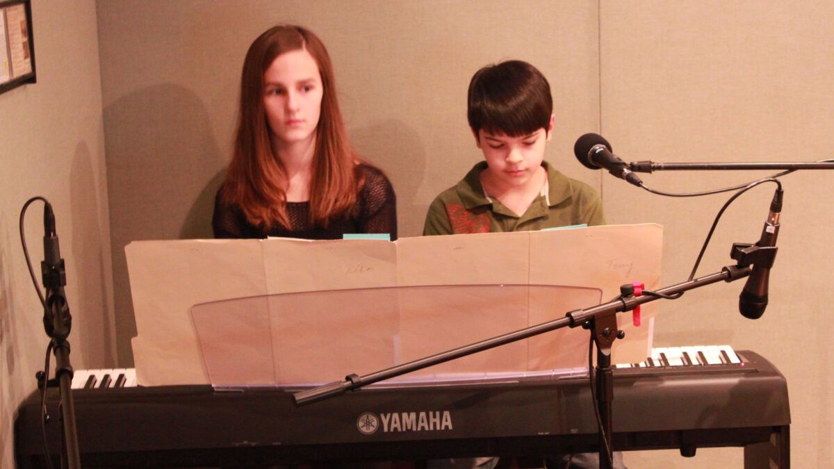 A teen and a youth playing a piano keyboard and looking at sheet music in a radio studio