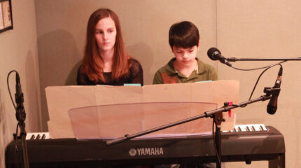 A teen and a youth playing a piano keyboard and looking at sheet music in a radio studio
