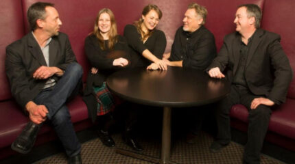 Five adults smiling and sitting on a large maroon couch around a round dark brown table