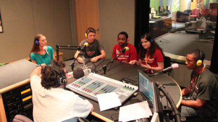 Five teens wearing headphones speaking into microphones being interviewed by an adult wearing headphones sitting behind a microphone and a mixer in a radio studio