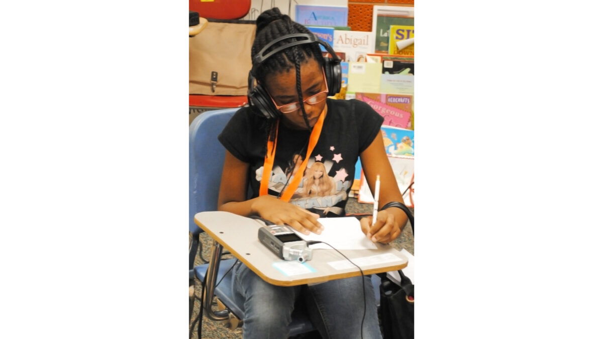 A youth wearing headphones connected to a portable recorder and writing on a piece of paper on a classroom desk
