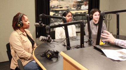 Two teens and an adult wearing headphones sitting behind a microphone being interviewed in a radio studio