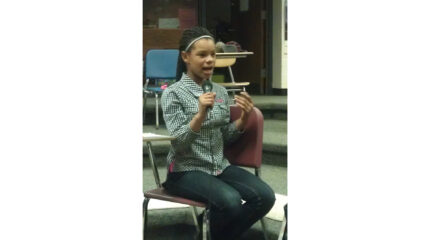 A youth speaking into a microphone while gesturing with their hands in a classroom