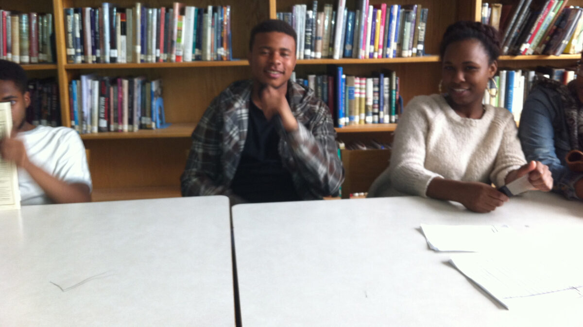 Three teens sitting in a library, two smiling at the camera