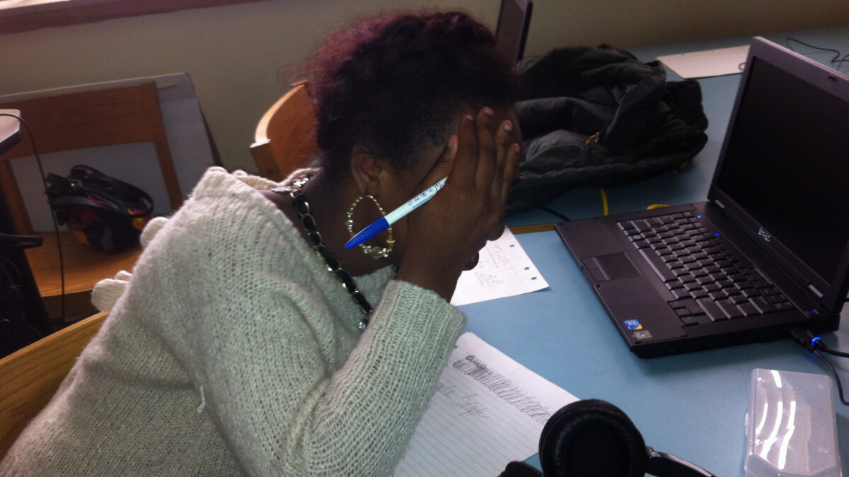 A teen covering their face while looking down at a piece of notebook paper and sitting behind a laptop computer