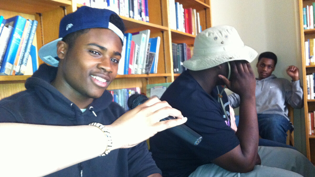 A hand holding a microphone for a teen sitting next to two other teens, one covering their face, in a library