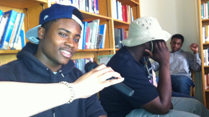 A hand holding a microphone for a teen sitting next to two other teens, one covering their face, in a library
